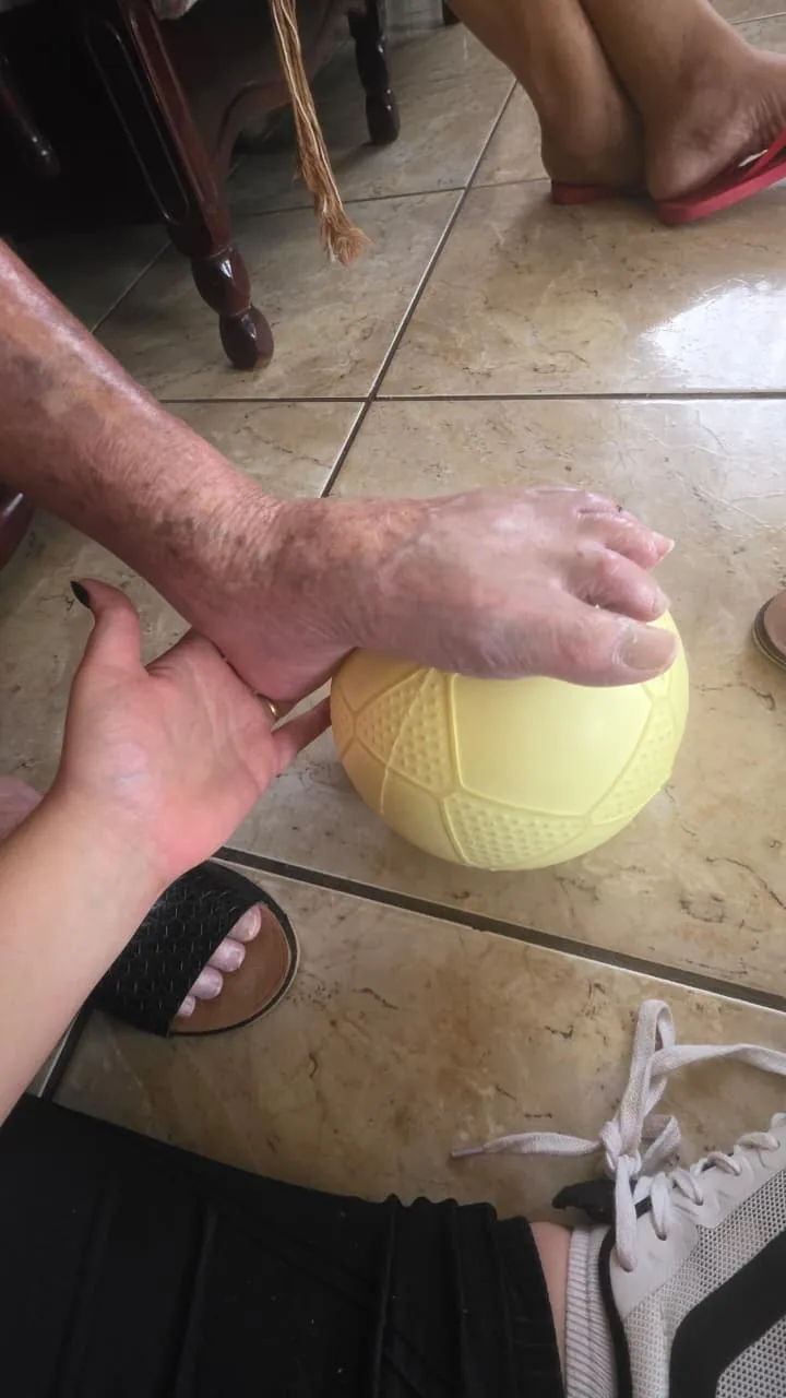 Fisioterapeuta apoiando a mão de uma pessoa idosa sobre uma bola amarela durante o atendimento.
